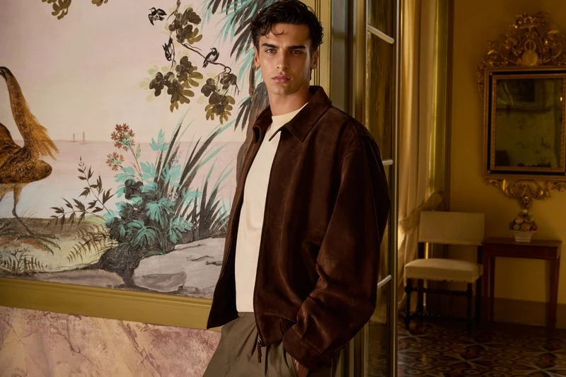A stylish man wearing a brown jacket stands indoors against a hand-painted wall in a well-lit room with elegant decor and natural light.