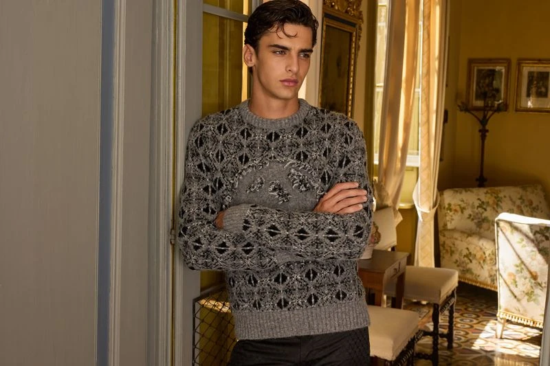 Indoor portrait of a dark-haired, light-skinned young man wearing a patterned sweater, standing with arms crossed, looking off to the side.