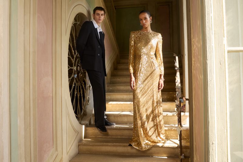 Two models posing on indoor stone stairs, a woman in a gold sequin gown and a man in a black suit.