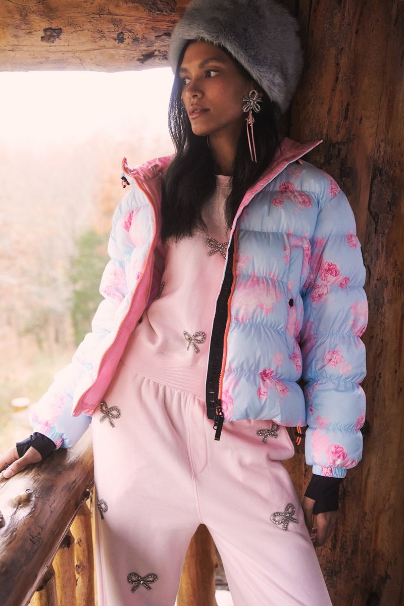 Brunette adult female wears floral-print puffer jacket, pink sweat suit, and fur hat near wooden balcony. Outdoor fashion photoshoot.