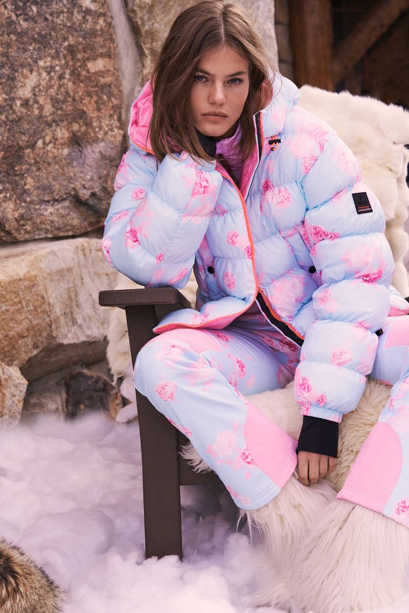 A woman sits in a chair wearing a puffy light blue and pink ski suit with a snowy mountain background on a sunny day.