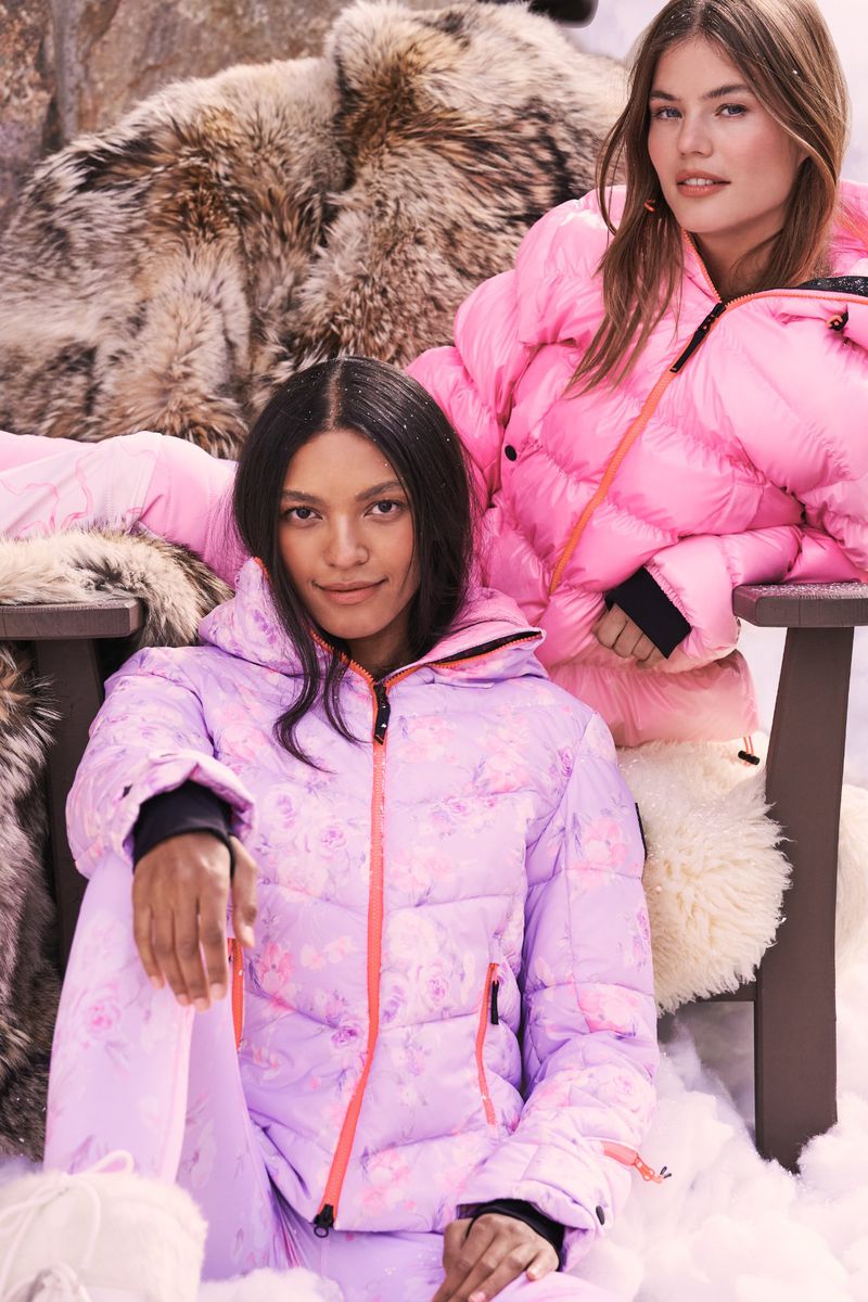 Two female fashion models pose in winter clothing. The models are wearing winter jackets and pants. Behind them is a fur throw.