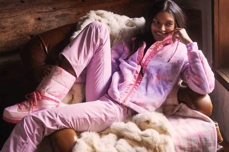 Smiling woman lounges in chair wearing pink snow pants, tie-dye jacket, and snow boots next to fur throw blanket indoors.