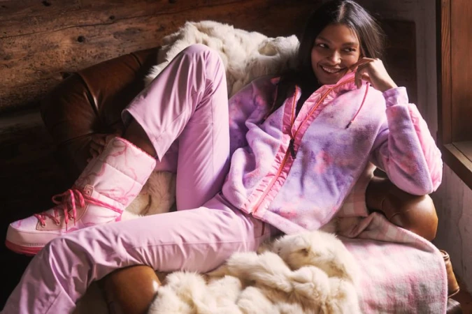 Smiling woman lounges in chair wearing pink snow pants, tie-dye jacket, and snow boots next to fur throw blanket indoors.