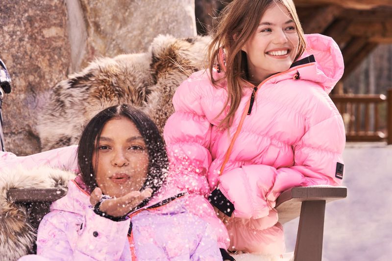 Two women in pink winter jackets sit outside on a wooden bench with rocks and trees visible in the background, blowing snow.