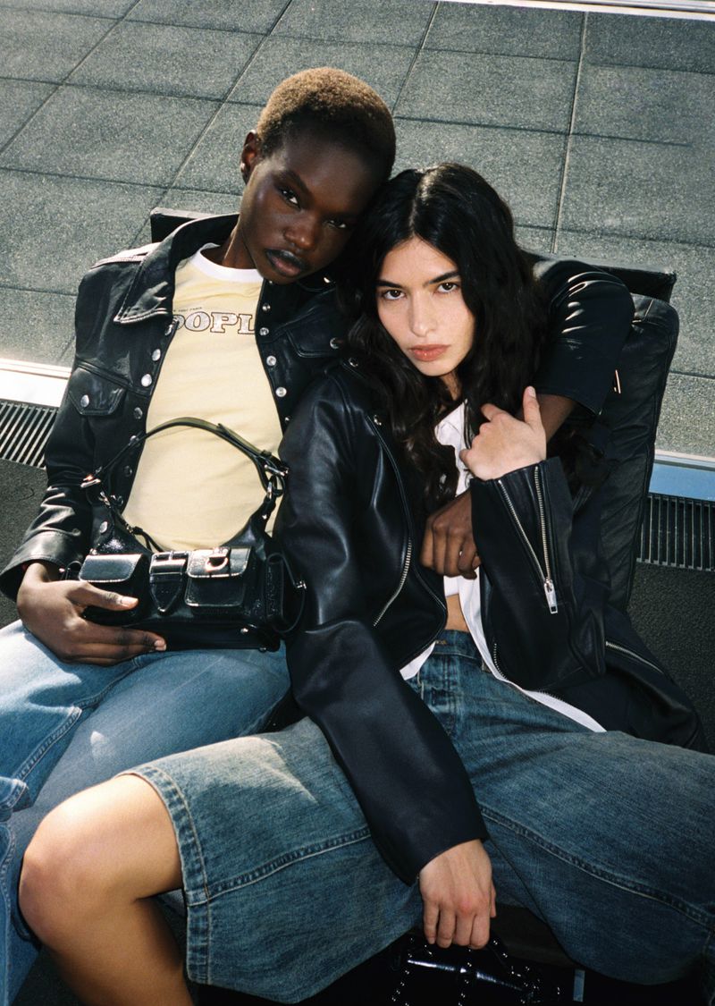Two women sit close together, wearing matching leather jackets and denim shorts, posing in an urban setting with a grey tiled background.
