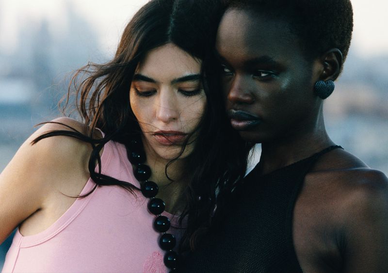 Two women pose closely together outdoors, one in pink and one in black, with a blurred cityscape background under soft lighting.