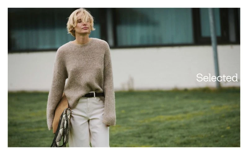 Adult woman in casual fashion with a sweater and white pants stands outside holding a satchel in front of a white building.