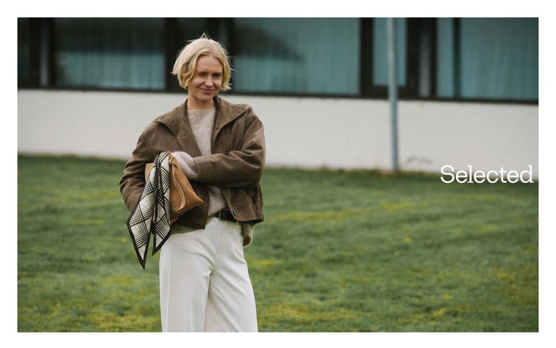 Woman in casual fashion stands on grass with a neutral expression, wearing a brown jacket and white pants in an outdoor setting.