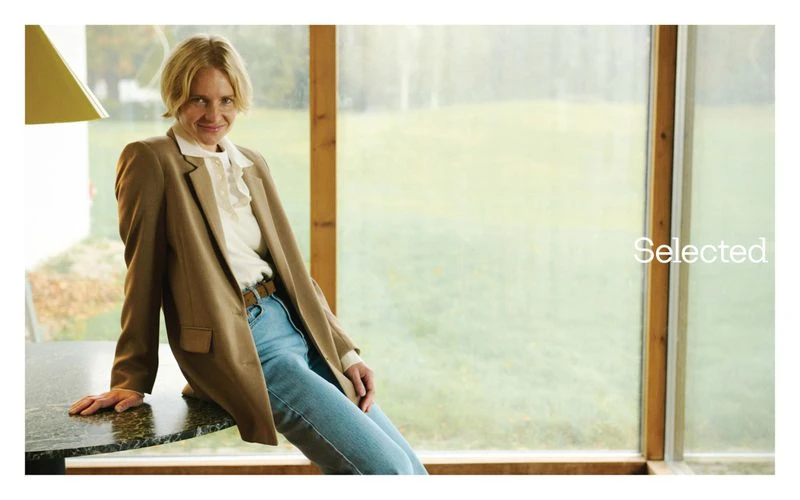 A fair-skinned adult woman with blonde hair leans against a table in a blazer, jeans, and white blouse in a bright, indoor location.