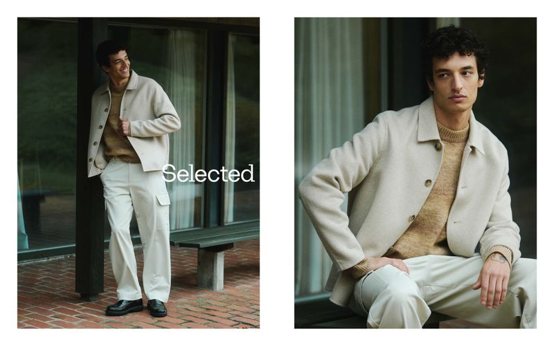 A brunette male model poses outdoors in a beige jacket, tan sweater, and off-white cargo pants for a fashion editorial shot in natural light.