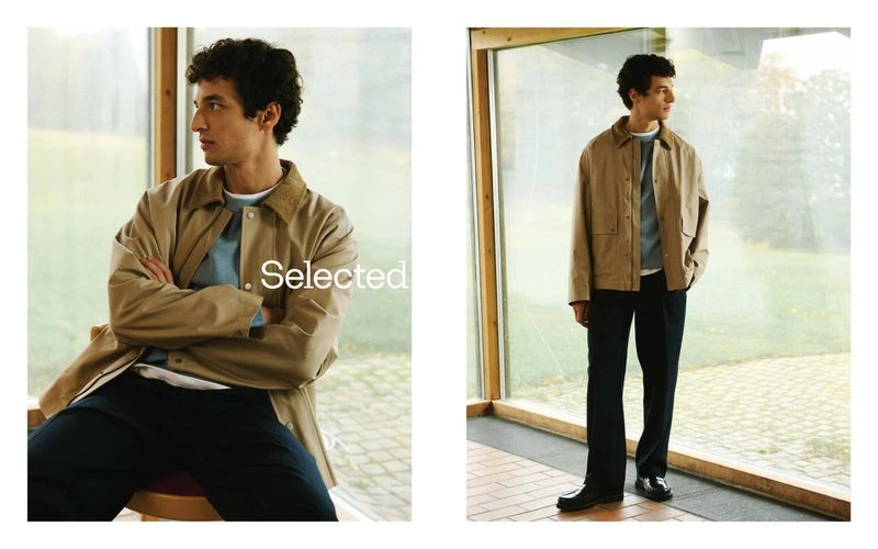 A young adult man in a beige jacket and dark pants poses in a casual setting with natural light and a clear window backdrop. Fashion.