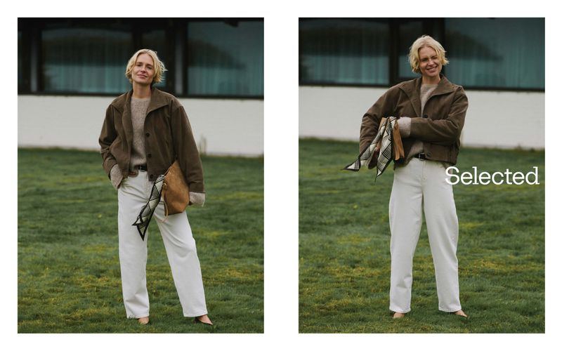 Blonde woman poses outdoors on grass wearing a brown jacket and white pants holding a tan bag in a lifestyle fashion shot.