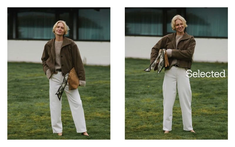 Blonde woman poses outdoors on grass wearing a brown jacket and white pants holding a tan bag in a lifestyle fashion shot.
