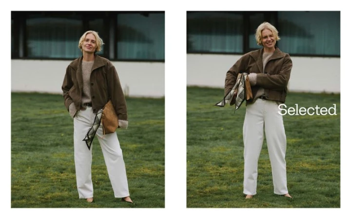 Blonde woman poses outdoors on grass wearing a brown jacket and white pants holding a tan bag in a lifestyle fashion shot.
