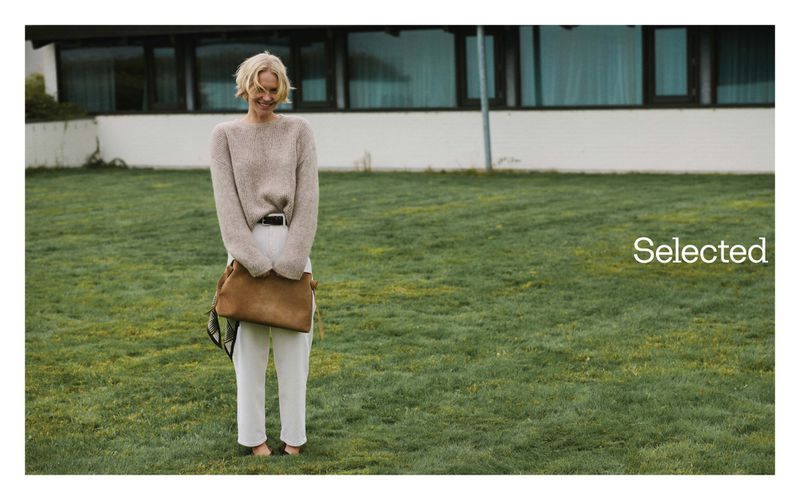 Blonde model standing on a lawn holding a brown leather bag, wearing a beige sweater and white trousers for Selected Q0 campaign.