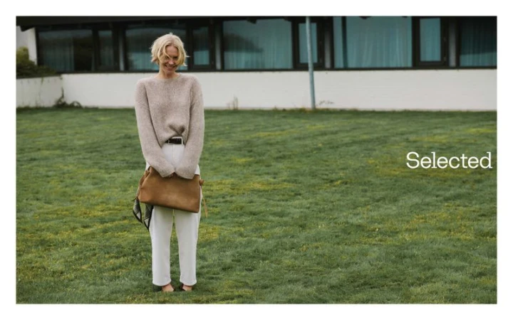 Blonde model standing on a lawn holding a brown leather bag, wearing a beige sweater and white trousers for Selected Q0 campaign.