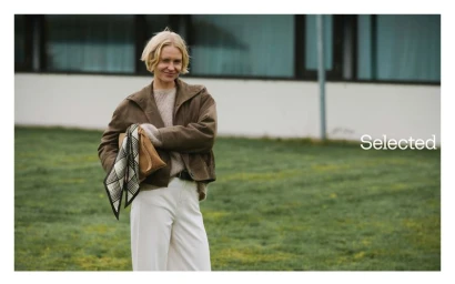 Blonde model wearing a brown jacket and white trousers outdoors with a patterned scarf and handbag for Selected.
