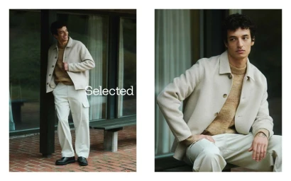 A man in a cream jacket, mustard knit sweater, and white trousers posing outside a modern building.