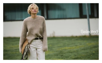 Model with blonde hair wearing a beige knit sweater and white trousers outdoors with a building in background