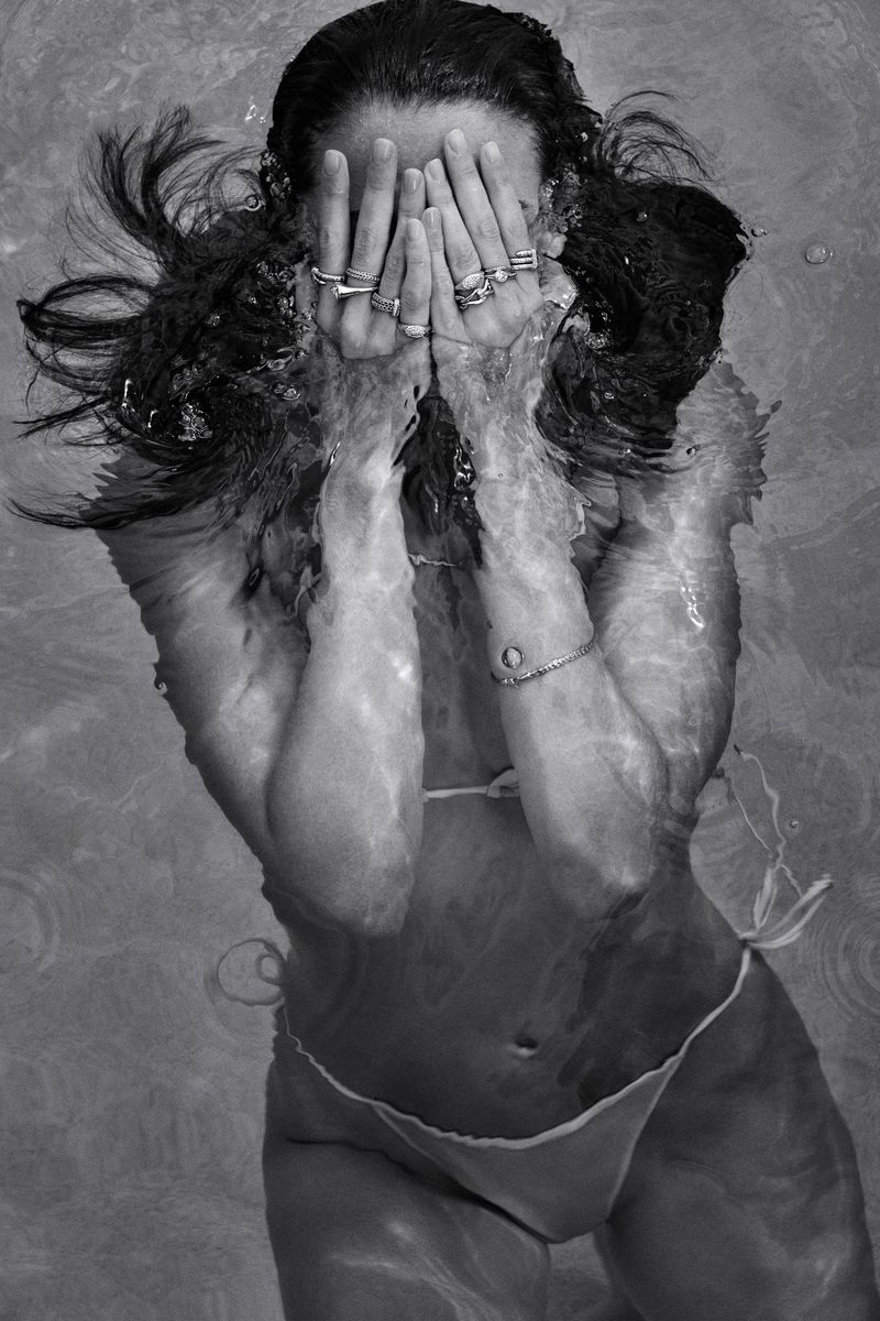 Monochromatic portrait of a woman in a bikini underwater, face partially hidden by her hands with jewelry, creating a surreal, ethereal aesthetic.