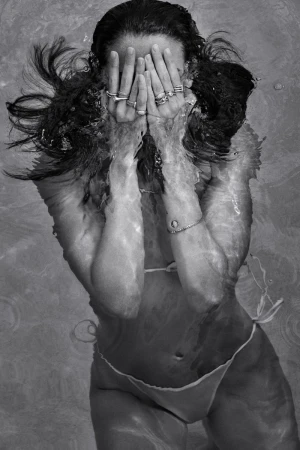 Monochromatic portrait of a woman in a bikini underwater, face partially hidden by her hands with jewelry, creating a surreal, ethereal aesthetic.