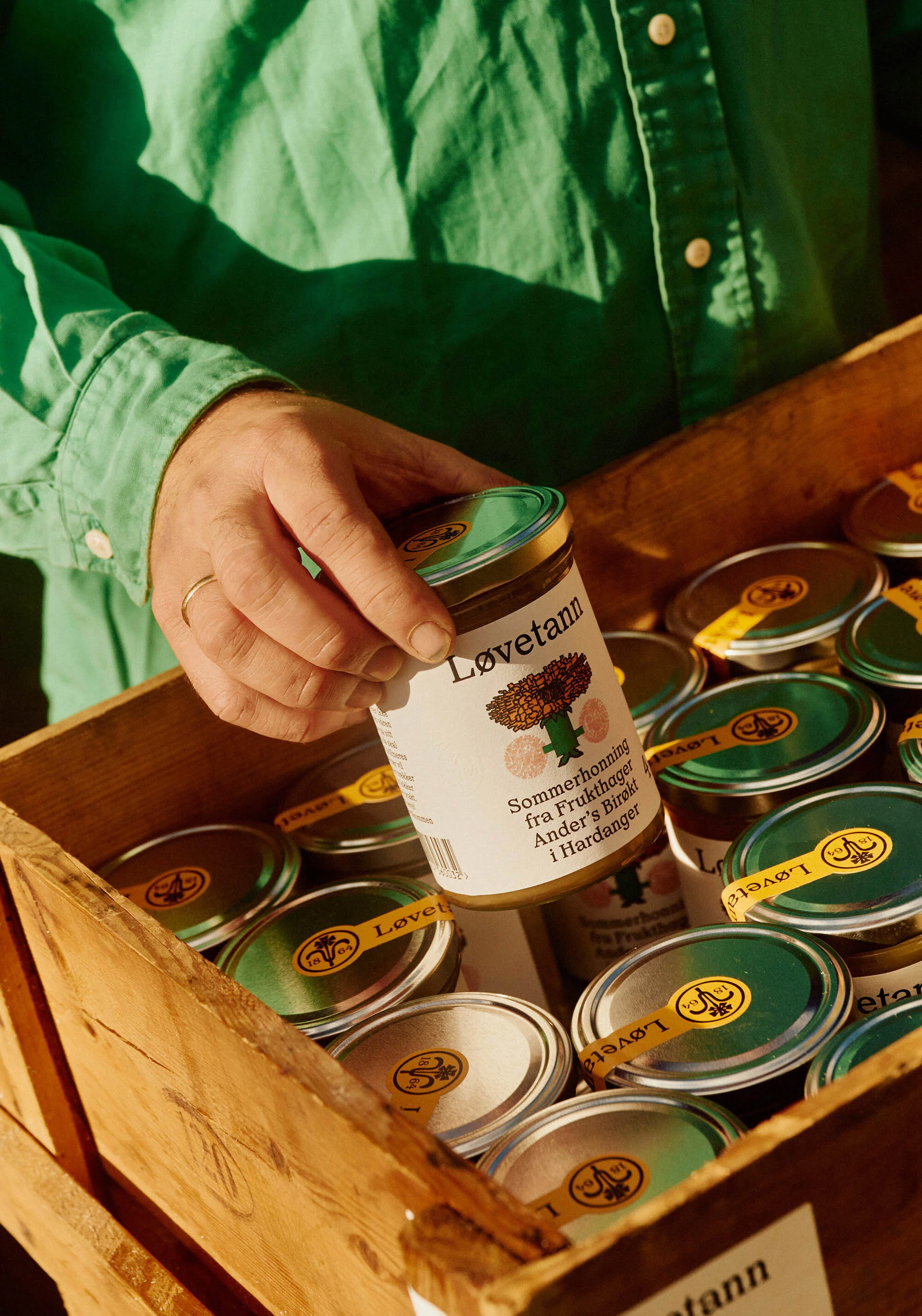 Person holds a jar of Lovetann honey in a wooden crate, filled with similar jars, labeled with green and gold lids. Outdoor setting.