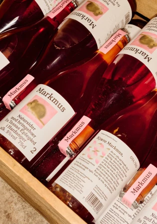 Several bottles of Markmus cider are arranged in a wooden container, showcasing their labels and the reddish hue of the liquid inside.