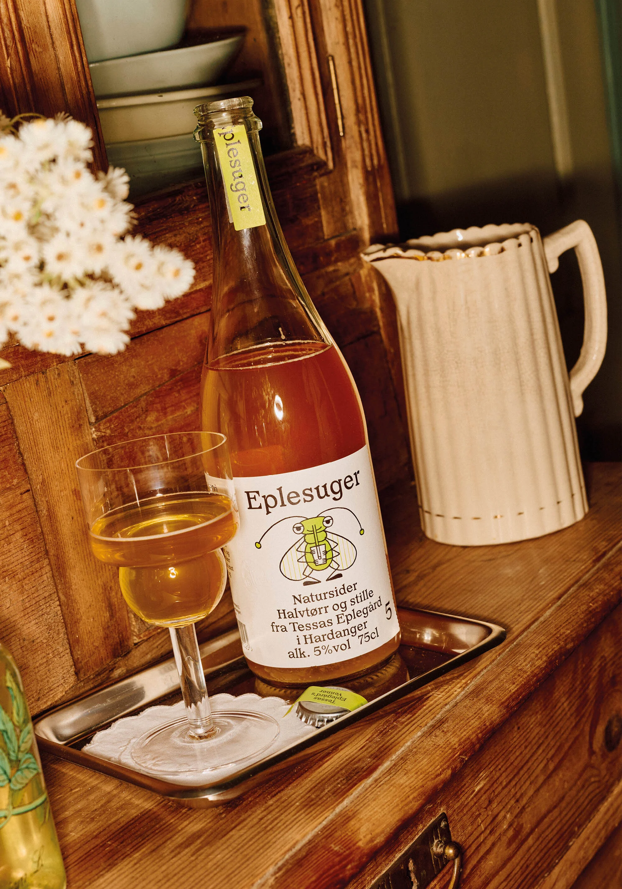 Bottle and glass of apple cider on a silver tray, accompanied by a pitcher, with rustic wooden furniture in the background.
