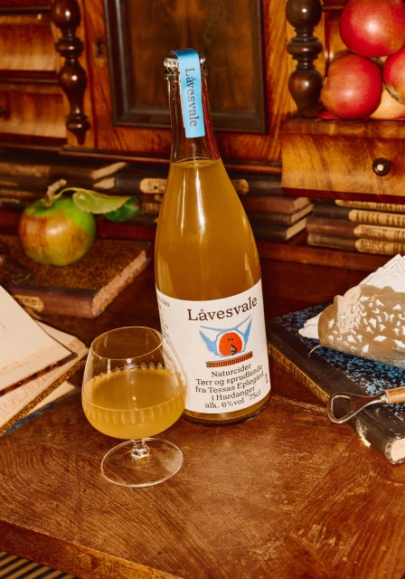 Bottle of Låvesvale cider with an apple and filled glass on a wooden table with books and other objects in the background.
