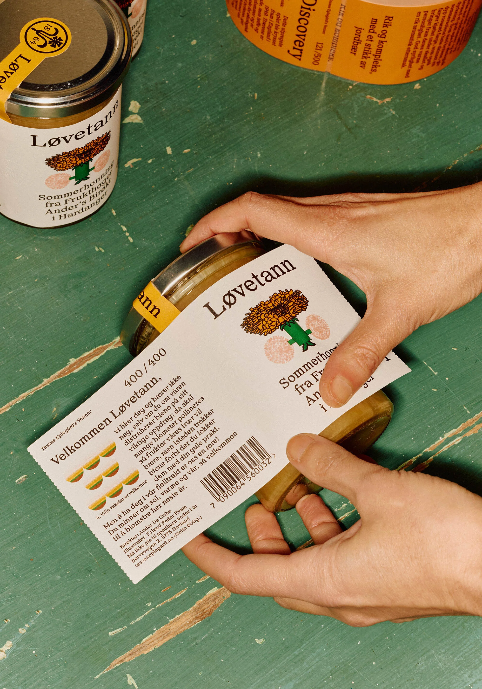 Close-up of hands labeling glass jars of Løvetann dandelion jelly, with a label featuring a dandelion graphic and Norwegian text.