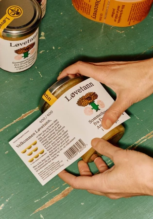 Close-up of hands labeling glass jars of Løvetann dandelion jelly, with a label featuring a dandelion graphic and Norwegian text.