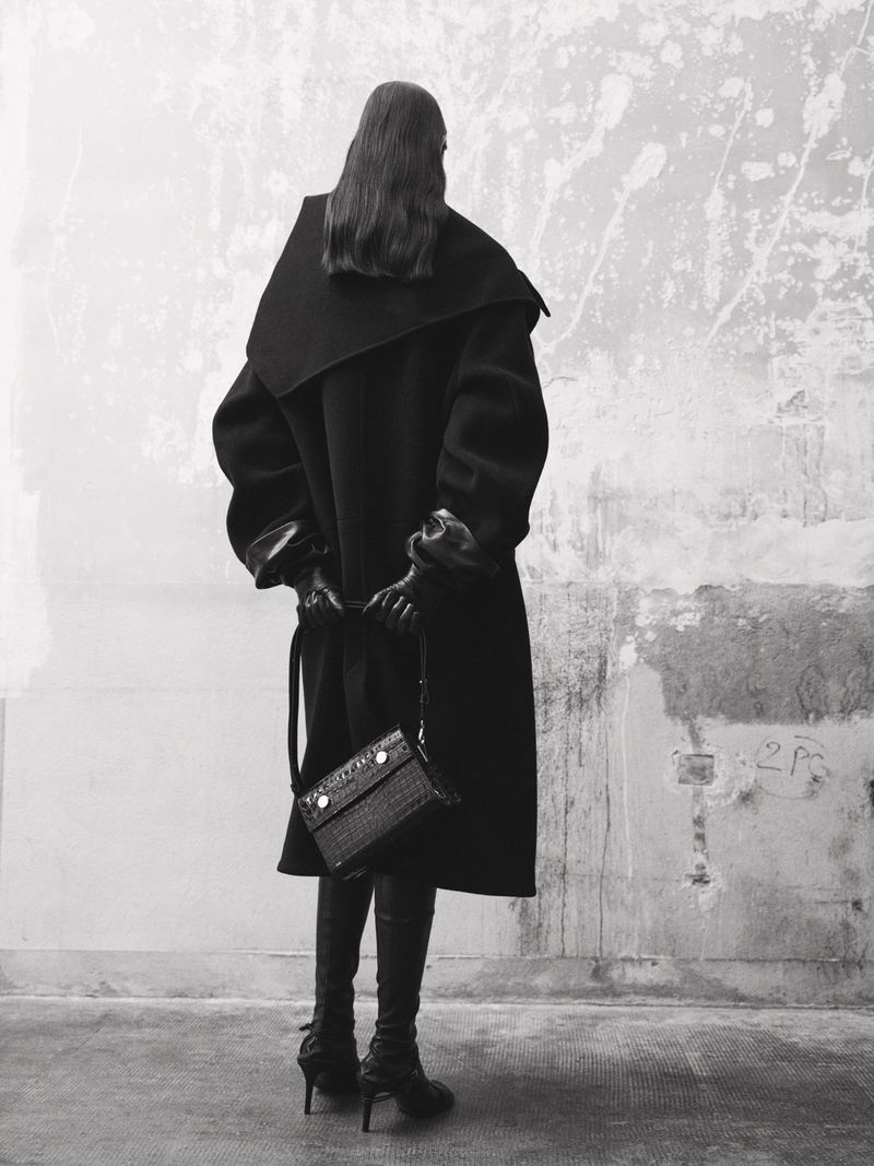 A woman stands with her back to the viewer, wearing a long black coat, knee-high boots, and carrying a textured black handbag against a mottled wall.