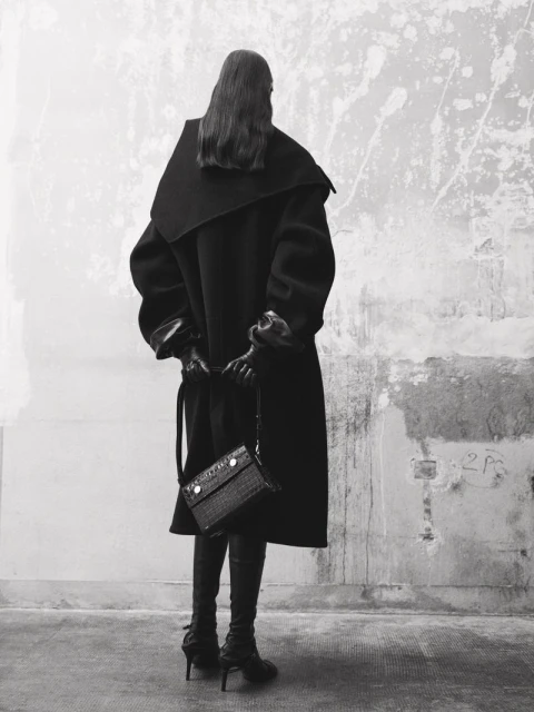 A woman stands with her back to the viewer, wearing a long black coat, knee-high boots, and carrying a textured black handbag against a mottled wall.