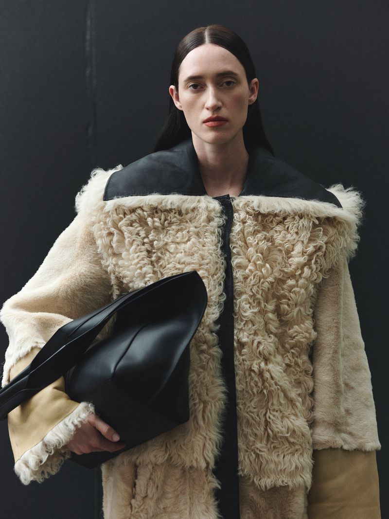 Elegant adult woman in fur coat and black bag poses against a dark background in a fashion-style photo. 