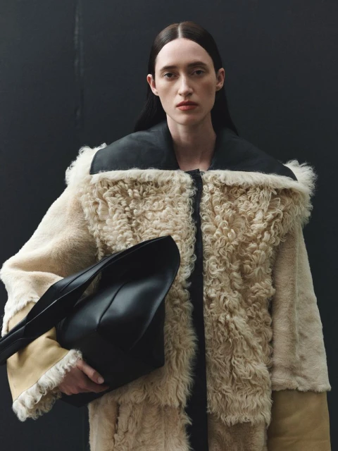 Elegant adult woman in fur coat and black bag poses against a dark background in a fashion-style photo. 