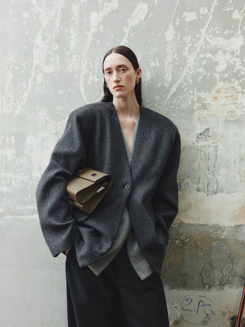 Fashion shot of a woman wearing a grey wool blazer and wide-leg pants, holding a brown bag, posed against a textured wall.