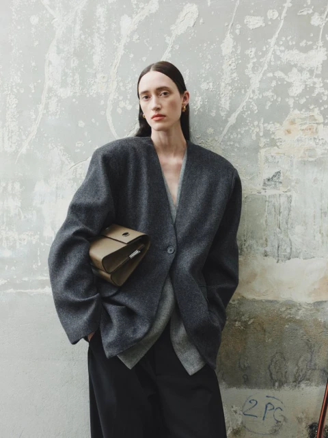 Fashion shot of a woman wearing a grey wool blazer and wide-leg pants, holding a brown bag, posed against a textured wall.