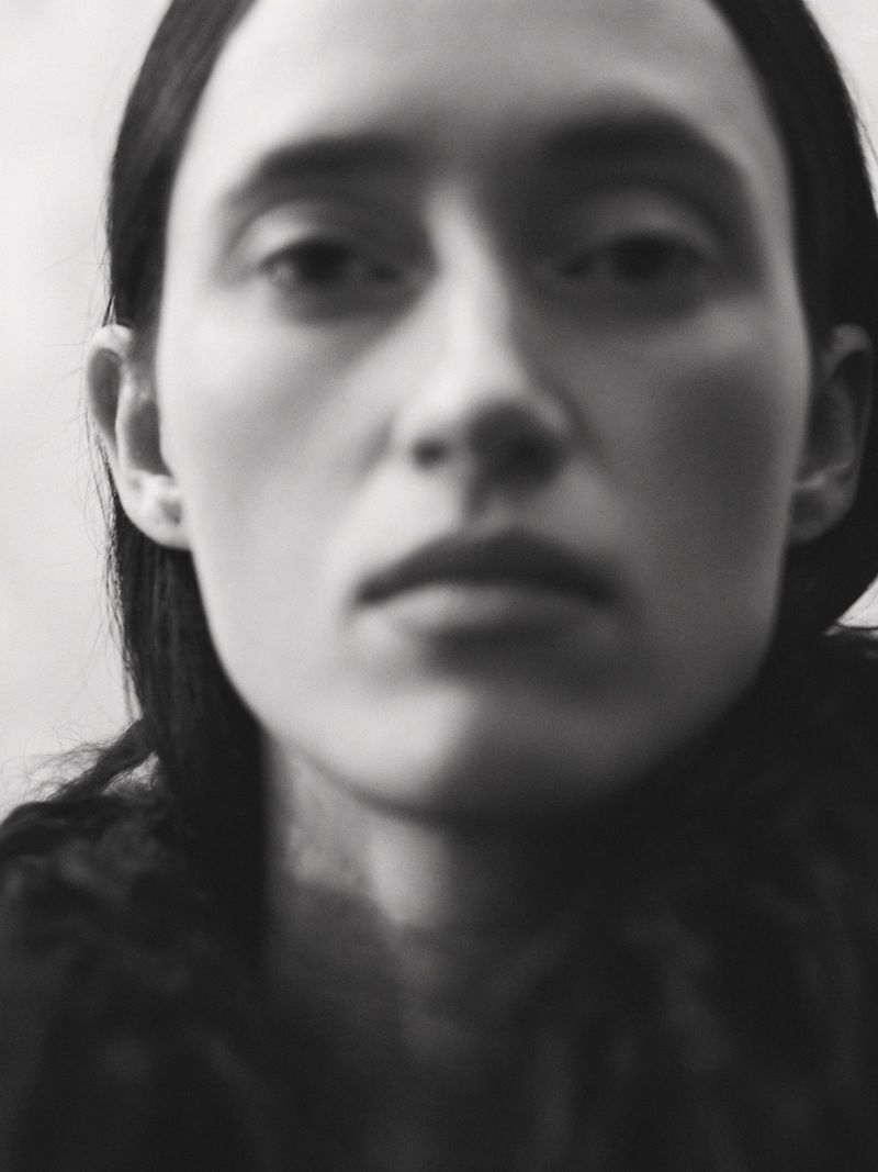 Black and white portrait of a young adult woman with straight dark hair, featuring a blurred background and shallow depth of field.