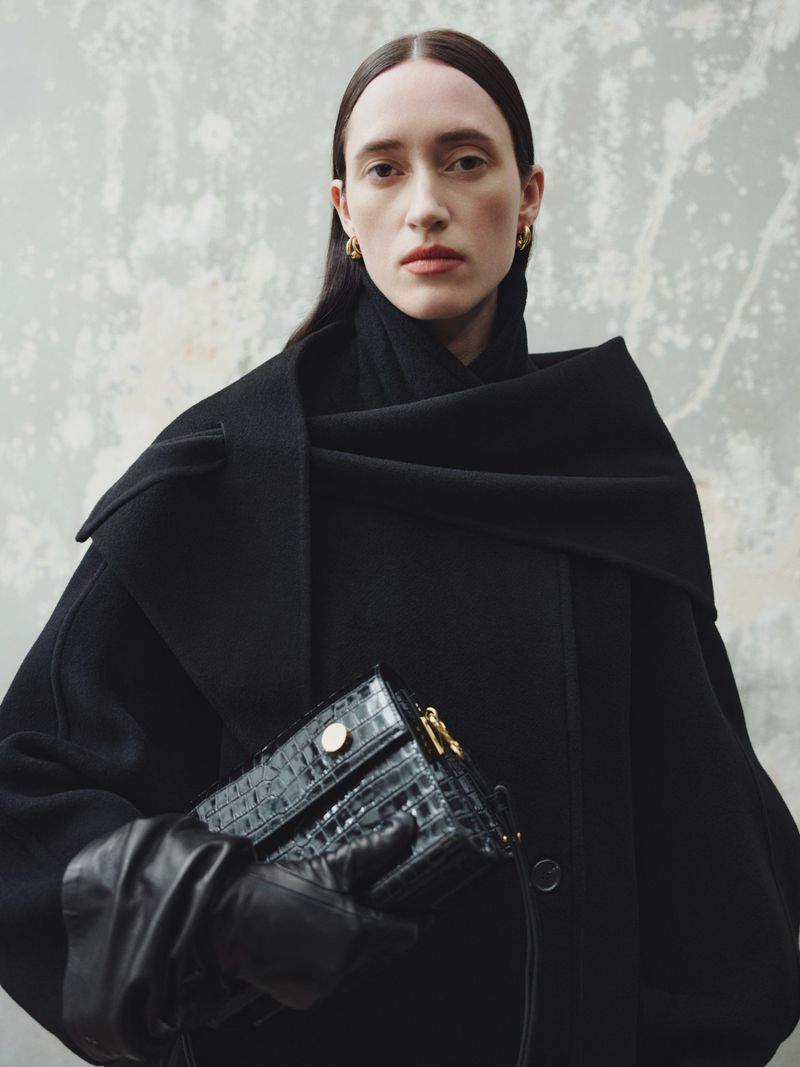 Elegant adult woman in a black coat holds a structured handbag against a textured grey backdrop in a studio fashion shot.
