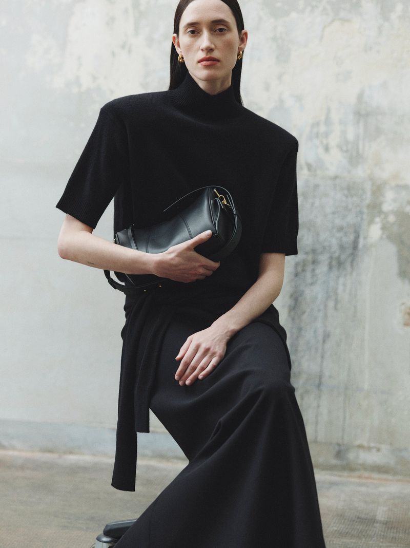 Model in black turtleneck and skirt holding a black leather handbag against a weathered wall background