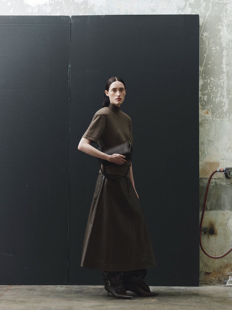 Woman in a brown dress and boots stands in front of a dark grey wall, holding a black clutch with a concrete background.