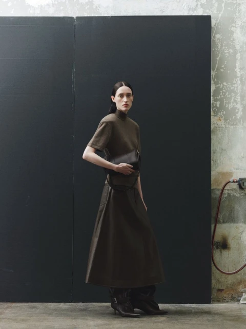Woman in a brown dress and boots stands in front of a dark grey wall, holding a black clutch with a concrete background.