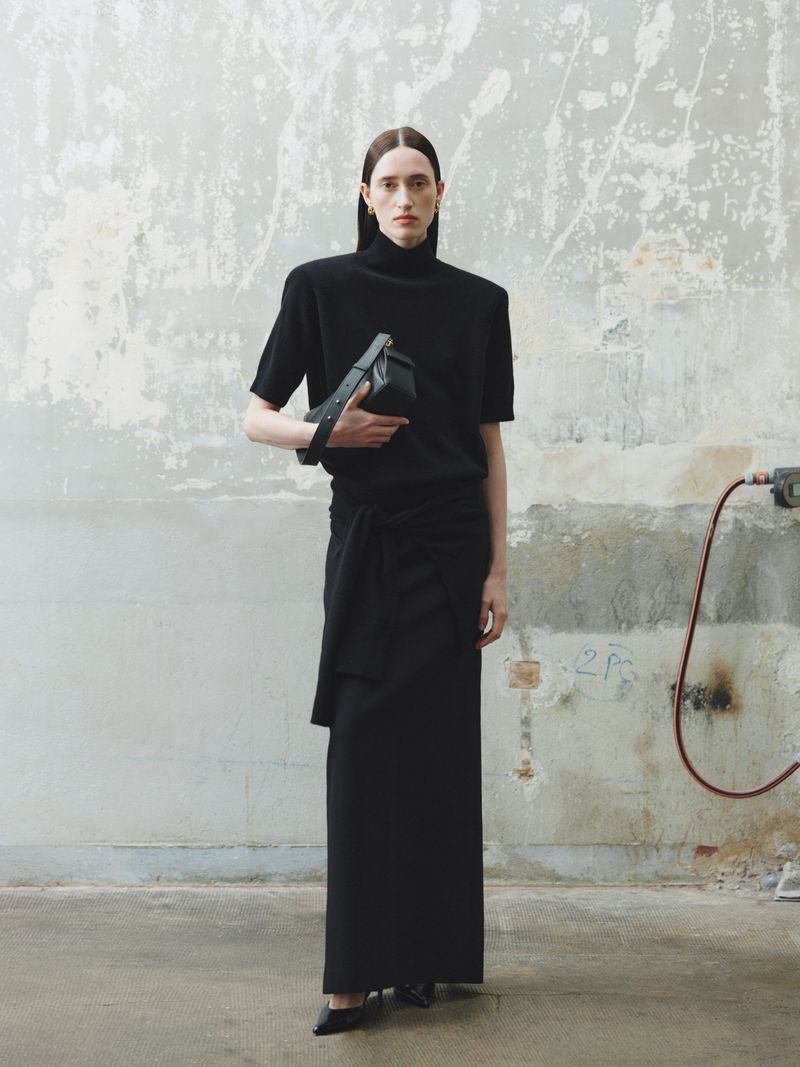 A woman poses in black skirt and top with a black purse, standing in front of a concrete wall in a fashion studio.