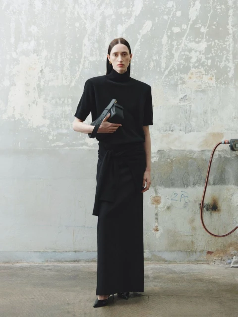 A woman poses in black skirt and top with a black purse, standing in front of a concrete wall in a fashion studio.