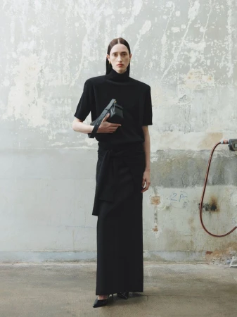 A woman poses in black skirt and top with a black purse, standing in front of a concrete wall in a fashion studio.