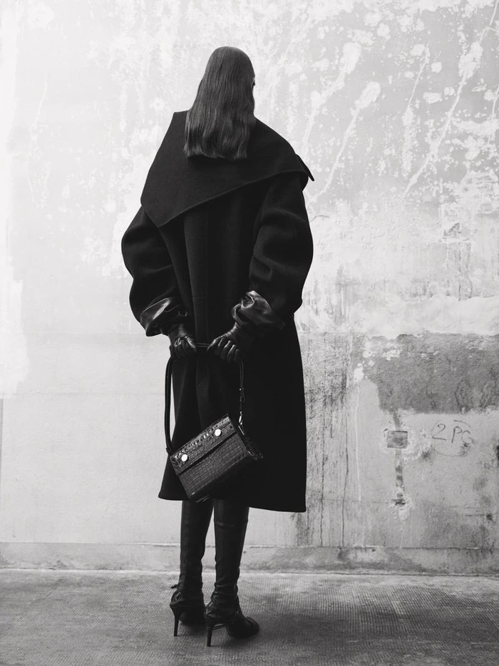 Model standing facing a weathered wall wearing a black coat and boots, holding a leather handbag from behind.