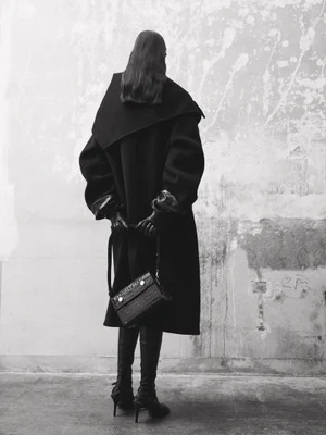Model standing facing a weathered wall wearing a black coat and boots, holding a leather handbag from behind.