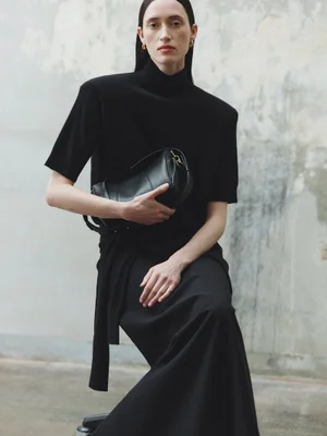 Model in black turtleneck and skirt holding a black leather handbag against a weathered wall background