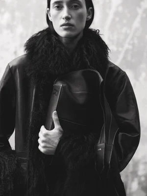 Model Helena Severin holding a dark leather bag while wearing a textured shearling collar coat in a black and white editorial portrait.